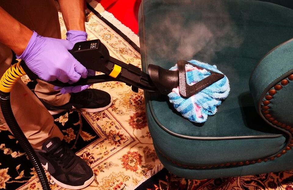 Person in purple gloves using steam cleaner on sofa during cleaning service in Dubai